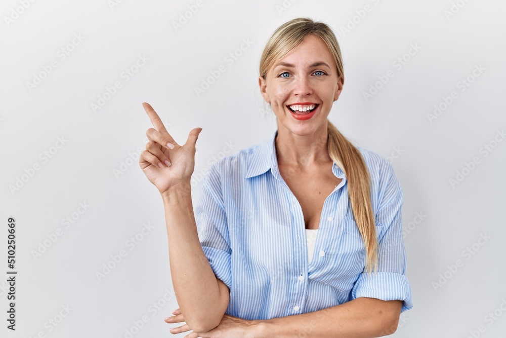 Poster beautiful blonde woman standing over white background with a big smile on face, pointing with hand a