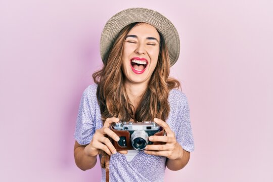 Young hispanic girl wearing summer hat holding vintage camera smiling and laughing hard out loud because funny crazy joke.
