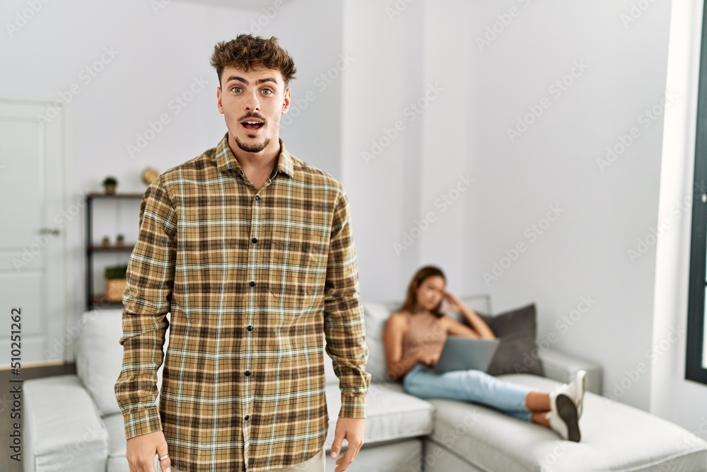 Poster Young handsome man at the living room with girlfriend scared and amazed with open mouth for surprise, disbelief face