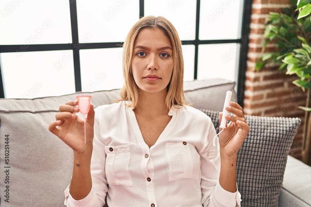 Sticker Young blonde woman holding menstrual cup and tampon relaxed with serious expression on face. simple and natural looking at the camera.