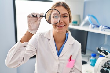 Young blonde woman working at scientist laboratory using magnifying glass looking positive and happy standing and smiling with a confident smile showing teeth