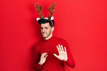 Young hispanic man wearing cute christmas reindeer horns disgusted expression, displeased and fearful doing disgust face because aversion reaction. with hands raised