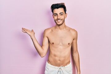 Fototapeta premium Young hispanic man wearing swimwear shirtless smiling cheerful presenting and pointing with palm of hand looking at the camera.