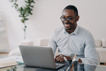 Smiling african american man wearing glasses working on business project at laptop at office desk