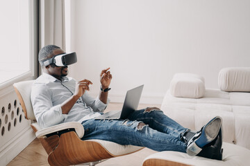 African businessman in VR glasses interact with virtual reality sit in armchair at laptop. High tech