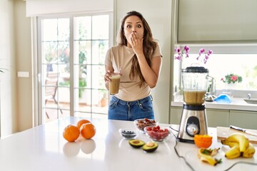Beautiful young brunette woman drinking glass of smoothie at the kitchen covering mouth with hand,...