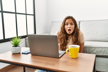 Beautiful hispanic woman using computer laptop at home scared and amazed with open mouth for surprise, disbelief face