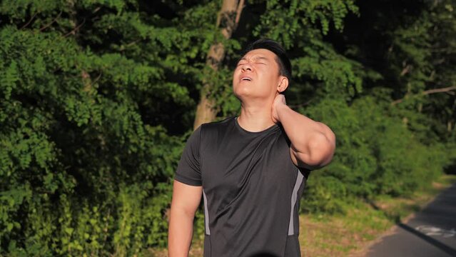 Asian Athlete running on the track feels sharp pain in the neck and stops. The runner feels pain in the cervical spine. Man holds neck with his hands. Male workout at sunset green trees background. 