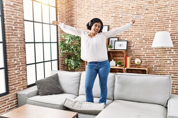 Young latin woman listening to music and dancing standing on sofa at home