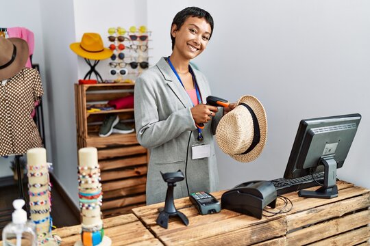 Young hispanic woman shopkeeper smiling confident scanning hat using bardcode reader at clothing store