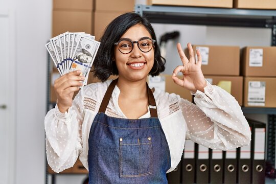 Young hispanic woman working at small business ecommerce holding dollars doing ok sign with fingers, smiling friendly gesturing excellent symbol