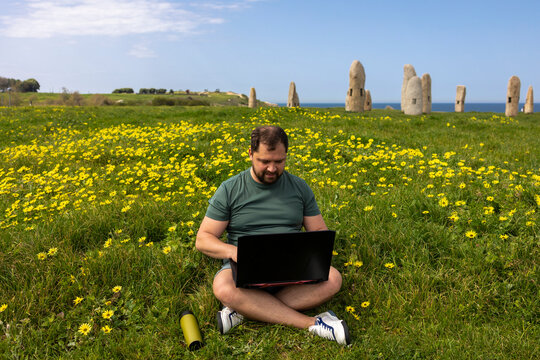 A man sits on a green grass with yellow flowers and works at a laptop. In the background is Stonehenge. Sunny warm day.