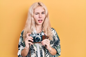 Beautiful caucasian woman with blond hair holding vintage camera winking looking at the camera with...