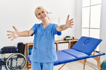 Beautiful caucasian physiotherapist woman working at pain recovery clinic looking at the camera smiling with open arms for hug. cheerful expression embracing happiness.