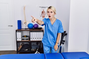 Young physiotherapist woman working at pain recovery clinic pointing with finger surprised ahead, open mouth amazed expression, something on the front