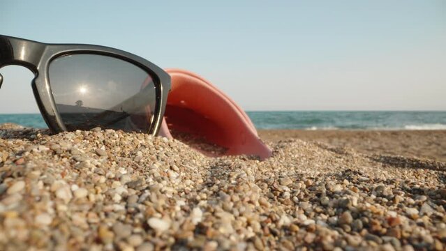 Sunglasses and pink flip flop lie in the sand against the background of the sea. The sun is reflected in glasses. Close-up. Laowa Probe.