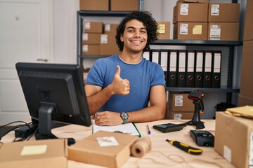 Hispanic man with curly hair working at small business ecommerce doing happy thumbs up gesture with hand. approving expression looking at the camera showing success.