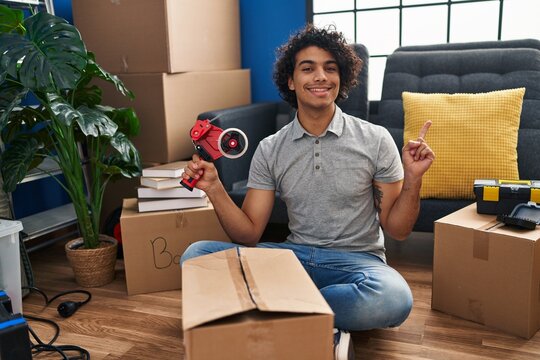 Hispanic man with curly hair moving to a new home closing cardboard box with tape smiling happy pointing with hand and finger to the side
