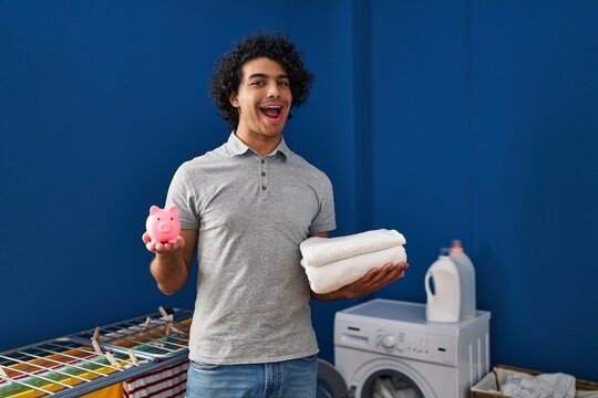 Hispanic man with curly hair holding clean towels and piggy bank celebrating crazy and amazed for success with open eyes screaming excited.