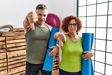 Middle age couple holding yoga mat looking unhappy and angry showing rejection and negative with...