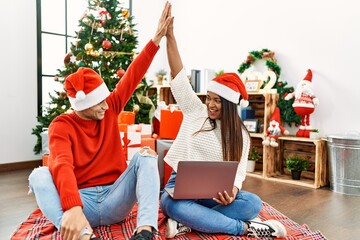 Young latin couple smiling happy and using laptop. Sitting on the floor high five by christmas tree...