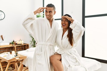 Young latin couple wearing towel standing at beauty center very happy and smiling looking far away with hand over head. searching concept.