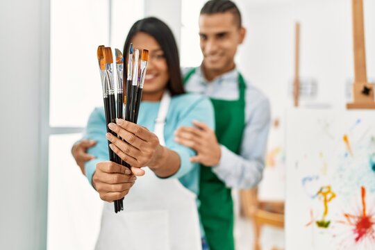 Young latin painter couple smiling happy holding paintbrushes at art studio
