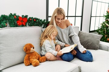 Mother and daughter reading book sitting by christmas tree at home