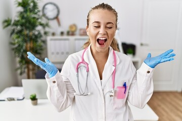 Young caucasian woman wearing doctor uniform and stethoscope at the clinic celebrating mad and crazy for success with arms raised and closed eyes screaming excited. winner concept