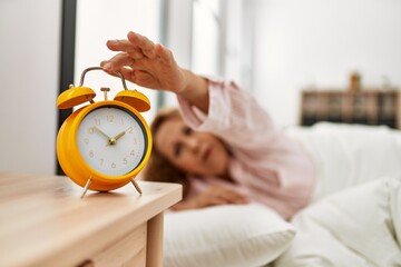 Middle age caucasian woman turning off alarm clock lying on the bed at bedroom.