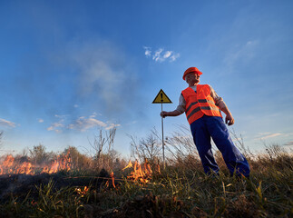 Firefighter ecologist fighting fire in field in evening. Man in orange vest and helmet near burning...