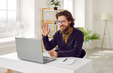 Happy handsome man sitting at desk, smiling and waving hello at laptop computer. Modern entrepreneur, remote corporate worker or teacher starts online work meeting or lesson via video call
