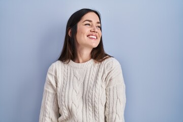 Young brunette woman standing over blue background looking away to side with smile on face, natural expression. laughing confident.