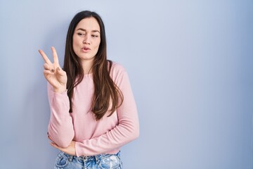 Young brunette woman standing over blue background smiling with happy face winking at the camera doing victory sign. number two.