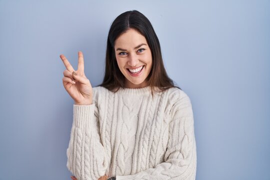 Young brunette woman standing over blue background smiling with happy face winking at the camera doing victory sign with fingers. number two.