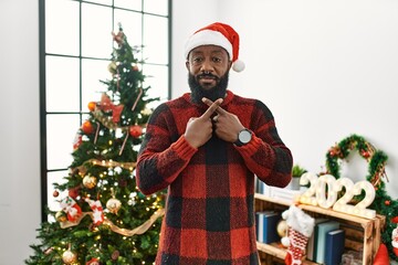 African american man wearing santa claus hat standing by christmas tree rejection expression crossing fingers doing negative sign