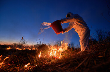 Research scientist fighting fire in field with blue night sky on background. Man in protective radiation suit and gas mask holding bucket and pouring water on burning grass with smoke.