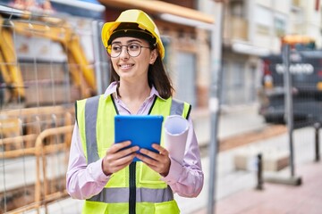Young hispanic woman architect using touchpad holding blueprints at street