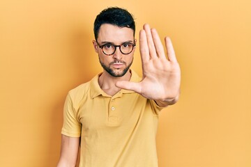Young hispanic man wearing casual clothes and glasses doing stop sing with palm of the hand. warning expression with negative and serious gesture on the face. - obrazy, fototapety, plakaty