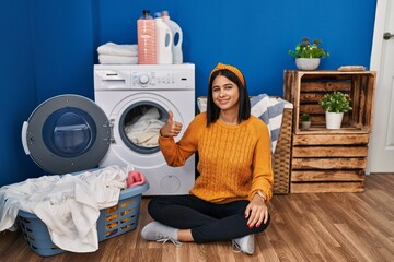 Young hispanic woman doing laundry doing happy thumbs up gesture with hand. approving expression looking at the camera showing success.