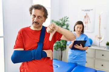 Fototapeta premium Southeast asian man wearing wearing arm on sling at rehabilitation clinic with angry face, negative sign showing dislike with thumbs down, rejection concept