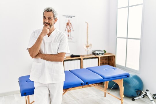 Middle age hispanic therapist man working at pain recovery clinic looking confident at the camera smiling with crossed arms and hand raised on chin. thinking positive.