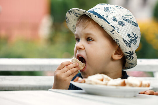Close up image of Caucasian baby boy with blonde hair and hat opening mouth going to bite crispy bread, having joyful facial expression.