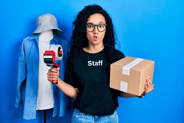 Young hispanic woman with curly hair wearing staff t shirt holding cardboard box making fish face...