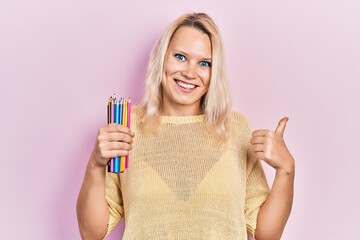 Beautiful caucasian blonde woman holding colored pencils pointing thumb up to the side smiling happy with open mouth