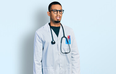 Young african american man wearing doctor uniform and stethoscope in shock face, looking skeptical and sarcastic, surprised with open mouth