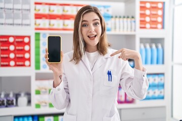 Blonde caucasian woman working at pharmacy drugstore showing smartphone screen pointing finger to one self smiling happy and proud