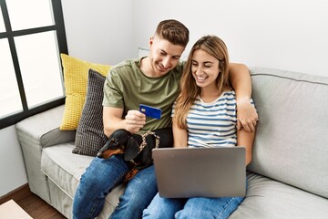 Young hispanic couple smiling happy using laptop and credit card sitting on the sofa  with dog at home.