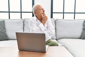 Senior man using laptop at home sitting on the sofa looking confident at the camera with smile with crossed arms and hand raised on chin. thinking positive. - Powered by Adobe