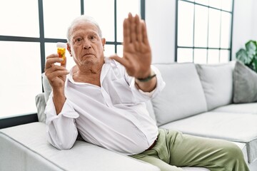 Senior man holding pills doing stop sing with palm of the hand. warning expression with negative and serious gesture on the face.